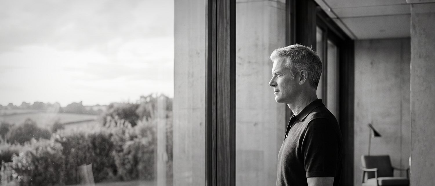 A man at a full-height window, looking out over open country